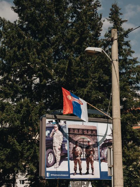 Plakat der Kfor am Strassenrand, davor weht die serbische Flagge