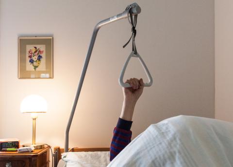 Symbolbild: Arm einer älteren Person hält sich am Bettgalgen eines Pflegebett