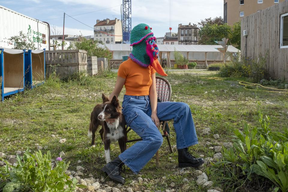 Fotoinszenierung: eine Person mit einer Mütze in Vogelform, welche den Kopf verdeckt, sitzt auf einem Stuhl und streichelt einen Hund