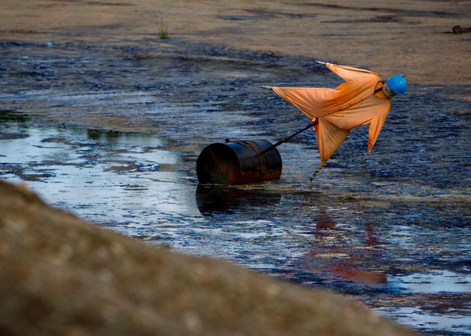 Symbolbild: Eine Vogelscheuche welche auf ein Ölfass montiert ist und in einem Ölsand-Becken schwimmt